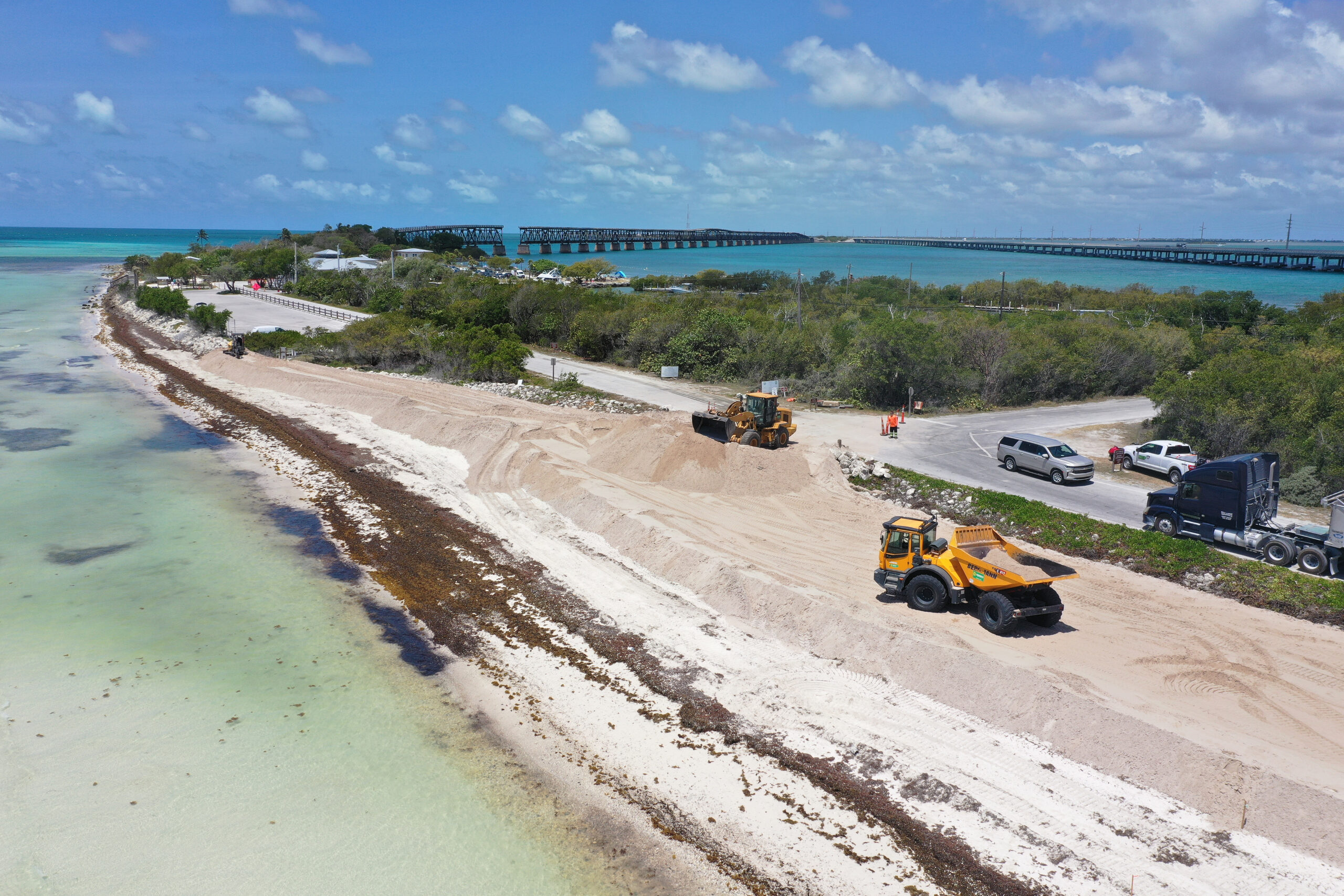 Heavy equipment rebuilding a beach along a coastal highway with bridges in the distance