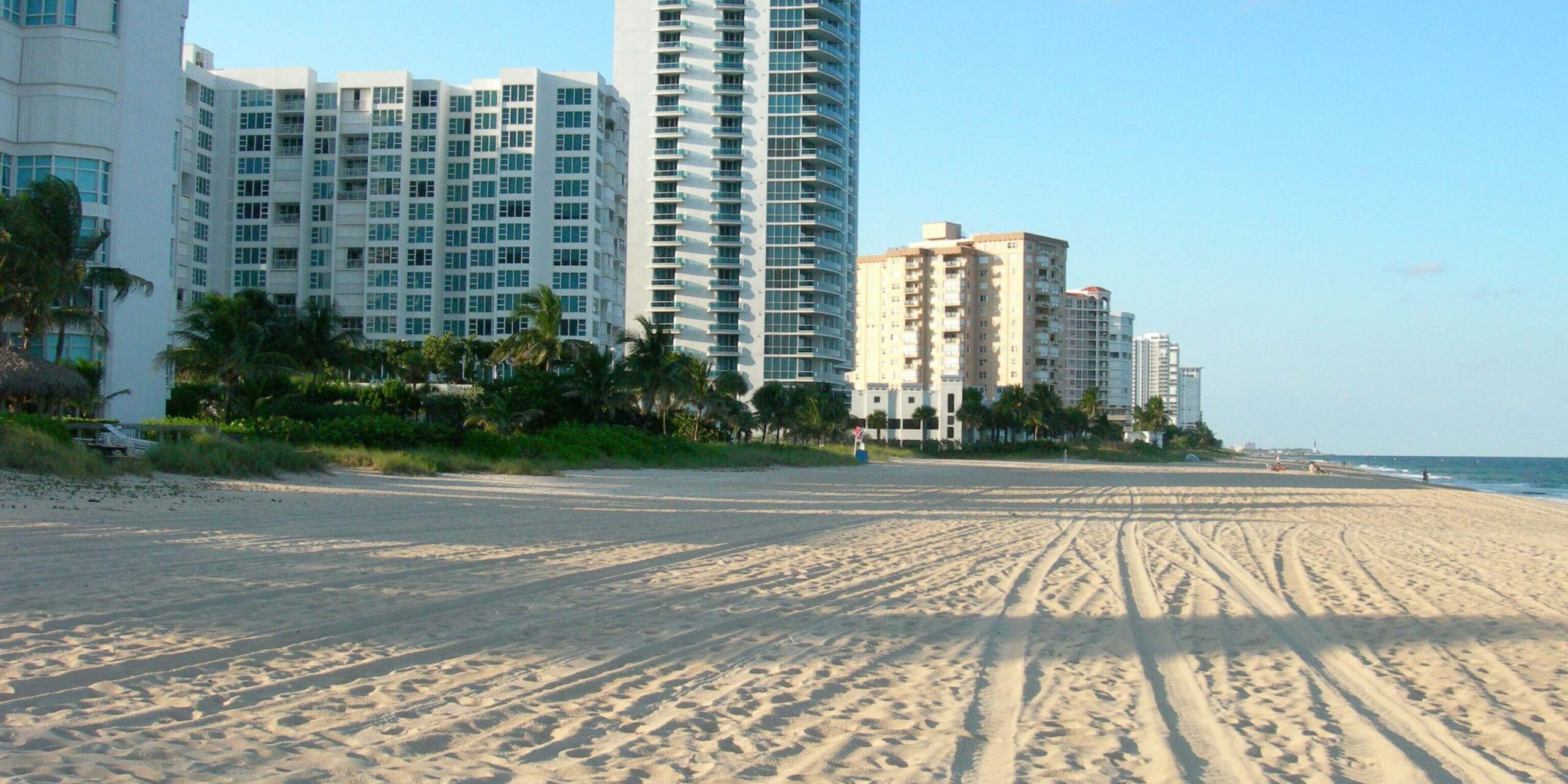 Sunny beach with footprints and tracks in the sand, bordered by a row of high-rise buildings under a clear blue sky